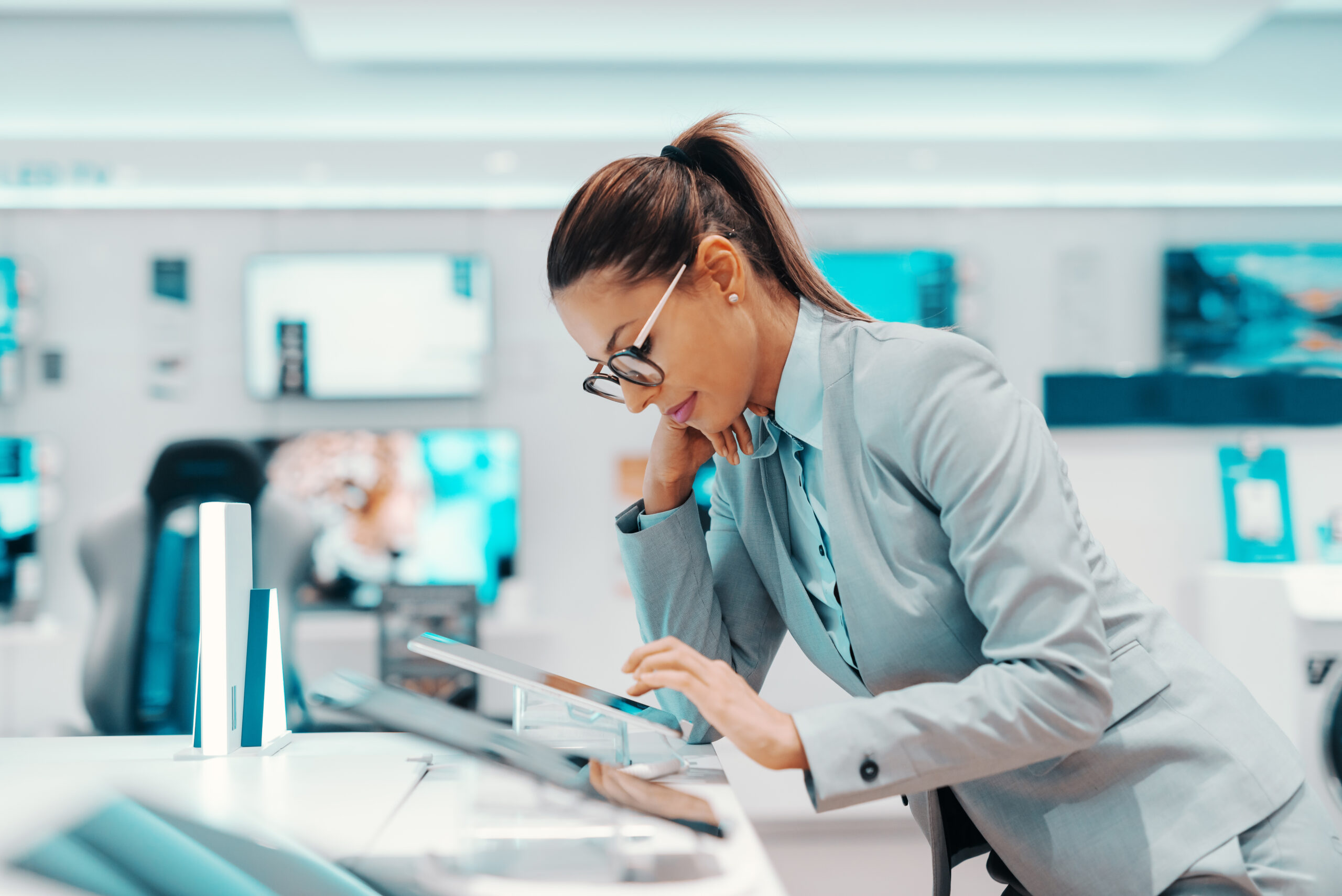 Pretty Caucasian brunette with ponytail dressed in formal wear and with eyeglasses leaning on counter and trying out tablet. Tech store interior.